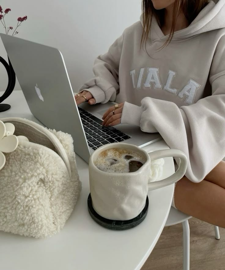 Cozy workspace with a laptop, coffee cup, and fluffy bag. Woman in a hoodie working from home, enhancing productivity and comfort.