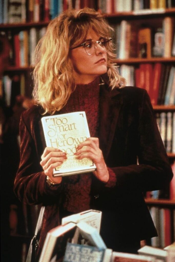 Woman holding a book titled Too Smart for Her Own Good in a library, wearing glasses and a brown jacket.