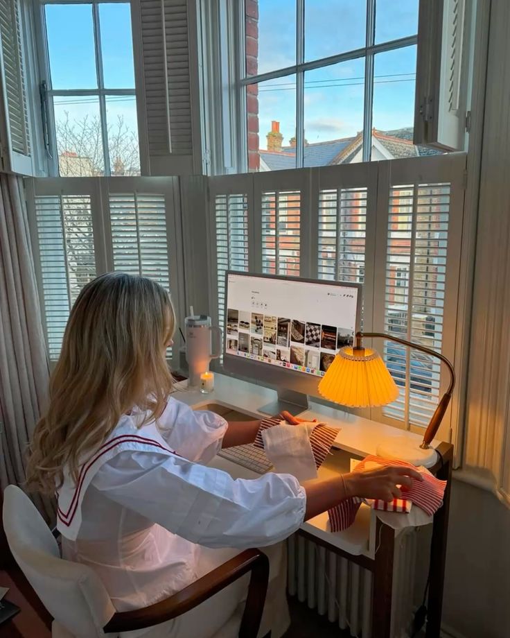 Person in white blouse sewing in cozy room, scenic view outside window, computer and lamp nearby. Ideal work-from-home setup.