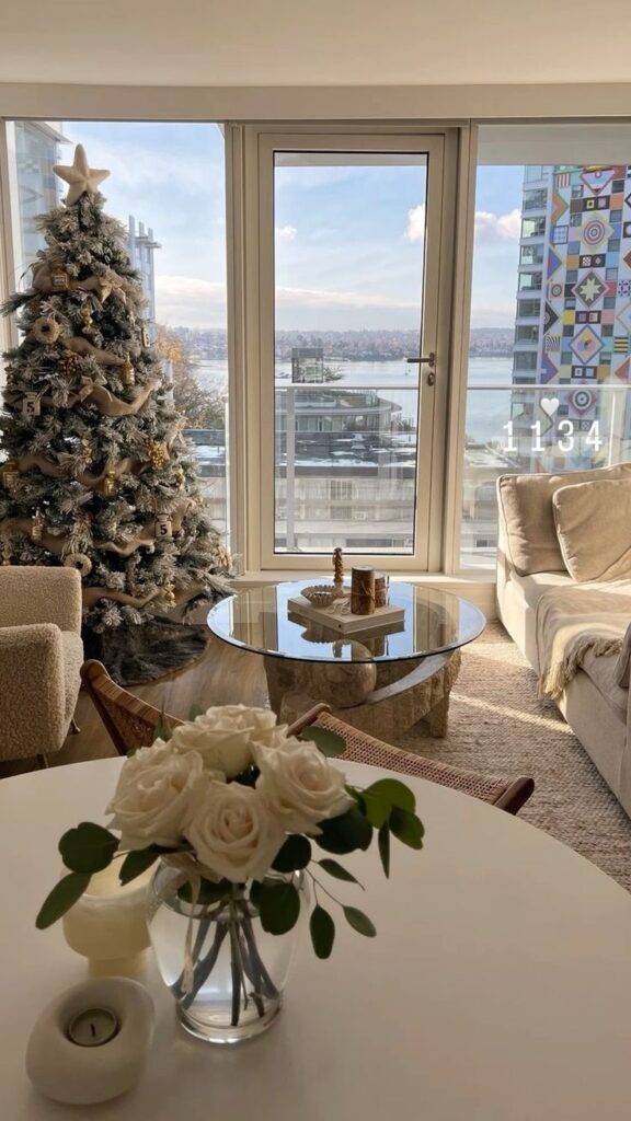 Cozy living room with a Christmas tree, white roses on a table, and a scenic window view of a winter landscape outside.