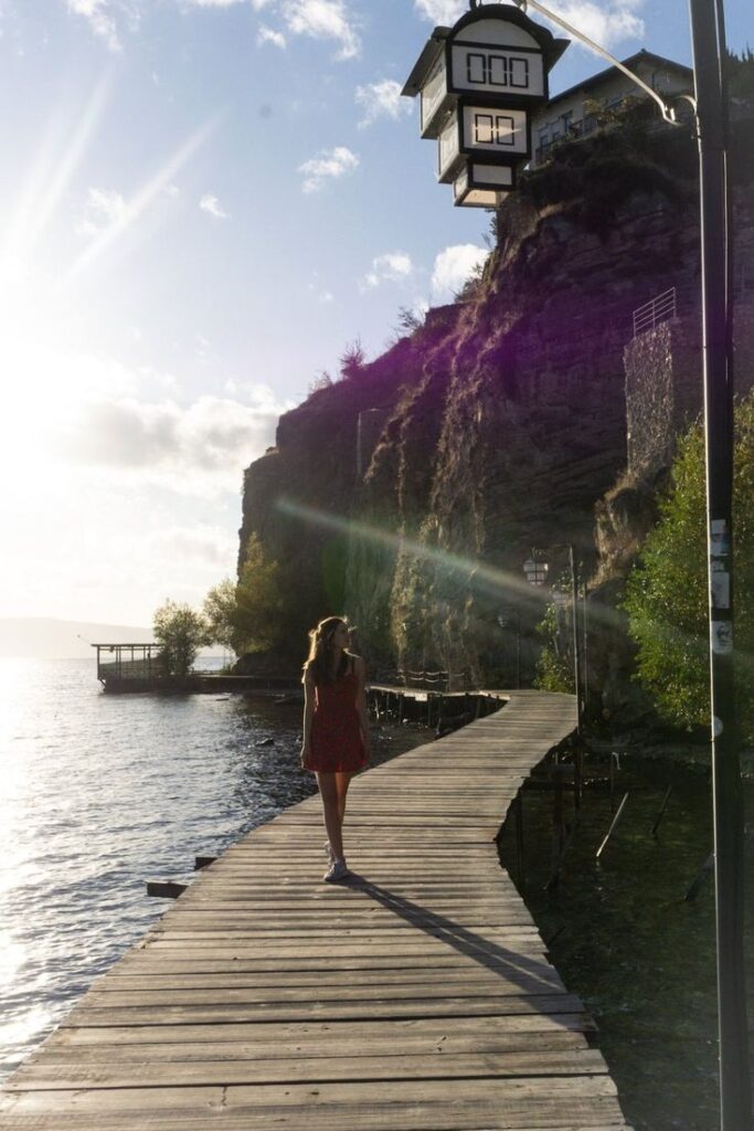 11 Things To Do In Ohrid North Macedonia - Handy Home Woman walking on a wooden pier by a cliffside at sunrise, overlooking a serene lake and lush greenery, with a unique lamp post above.