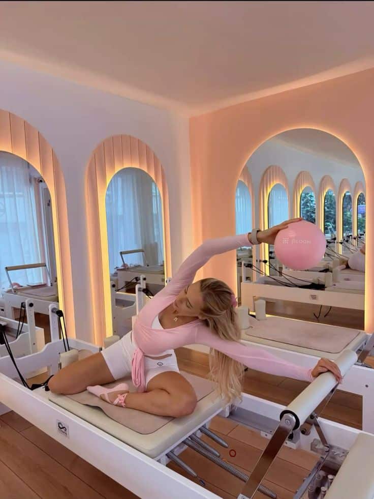 Woman doing pilates on a reformer exercise machine, stretching with a pink fitness ball in a serene studio with arched mirrors.