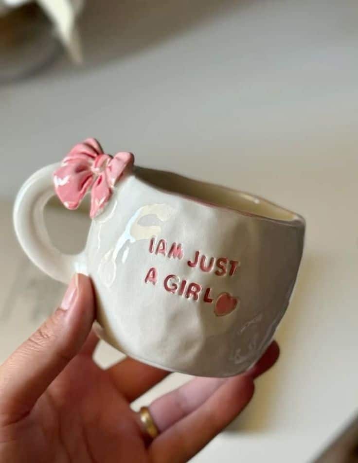 Ceramic coffee cup with pink bow and text I AM JUST A GIRL held in hand. Cute and whimsical design.