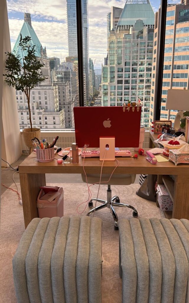 Colorful desk setup with a red computer, stationery, and a city view from high-rise office windows. Cozy and creative workspace.