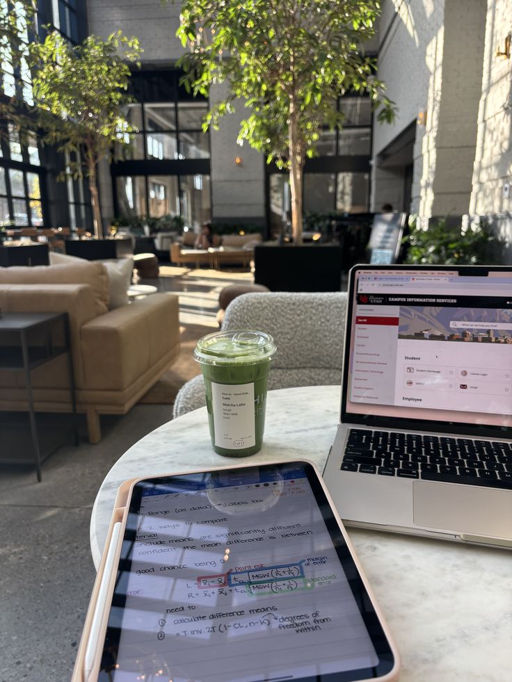 Cozy café work setup with a laptop, tablet, and green matcha drink; bright interior with plants and natural light.