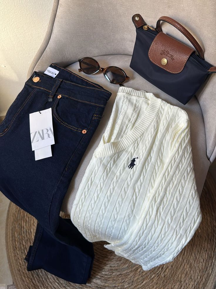 Folded jeans, white sweater, sunglasses, and a stylish handbag on a chair, showcasing a chic casual outfit arrangement.