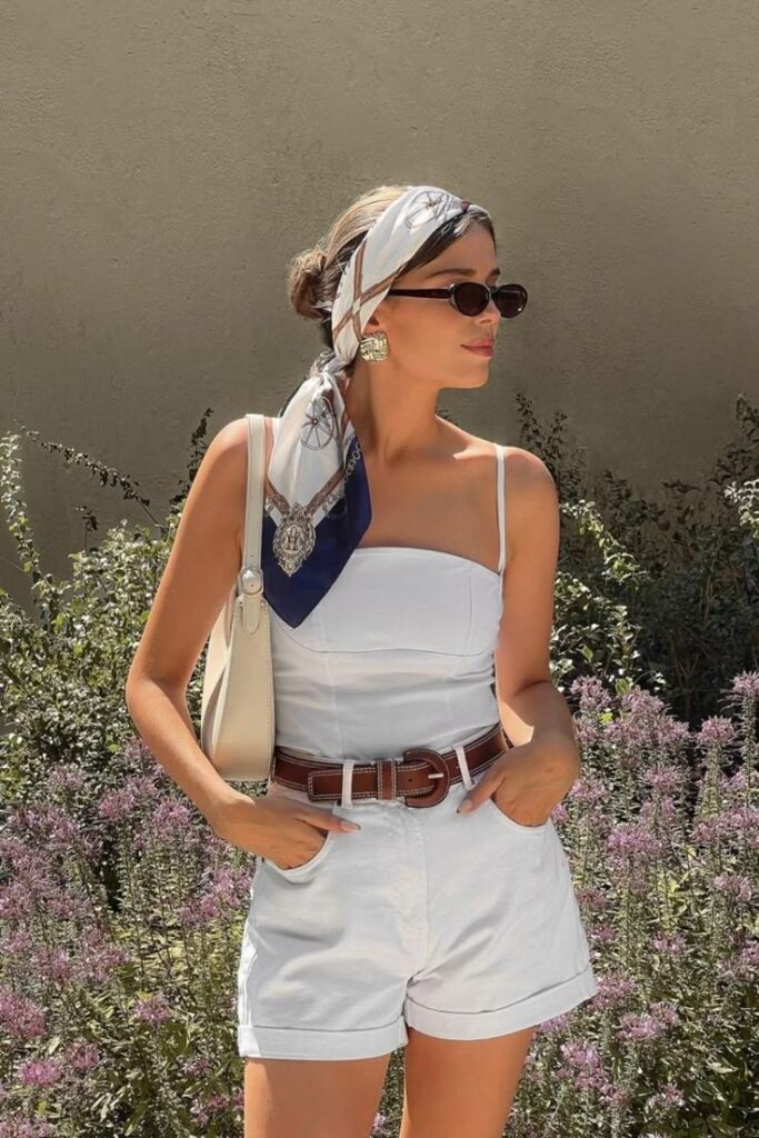 Stylish woman in white outfit with a scarf and sunglasses, standing in a garden. Casual summer fashion and accessories.