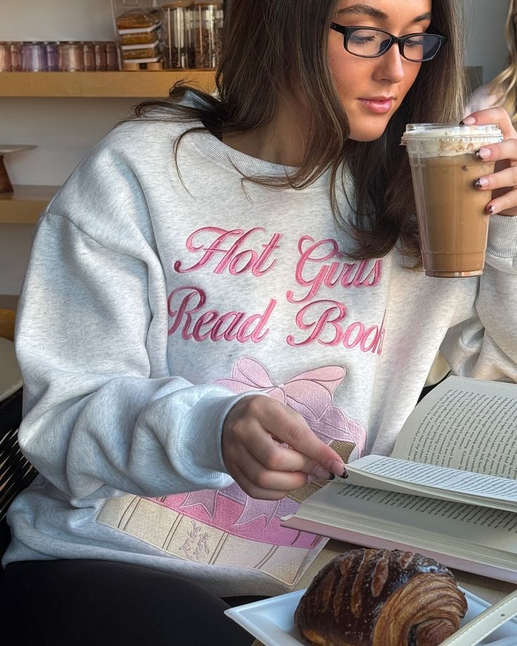 Woman in glasses reads a book, enjoying iced coffee and pastry. Sweatshirt reads Hot Girls Read Books. Cozy setting.