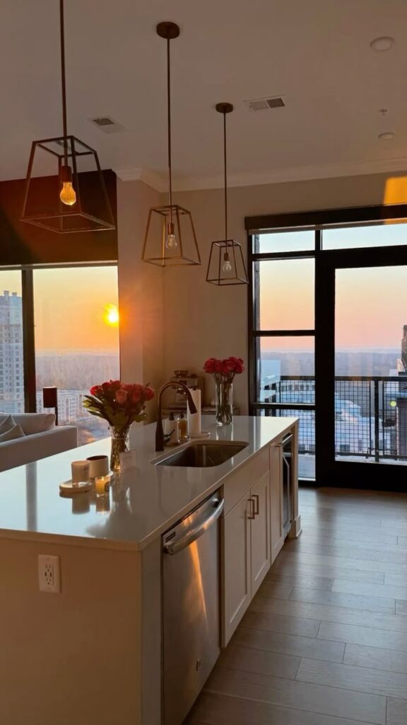 Modern kitchen with island and pendant lights, city view, and sunset through large windows, creating a warm and inviting atmosphere.