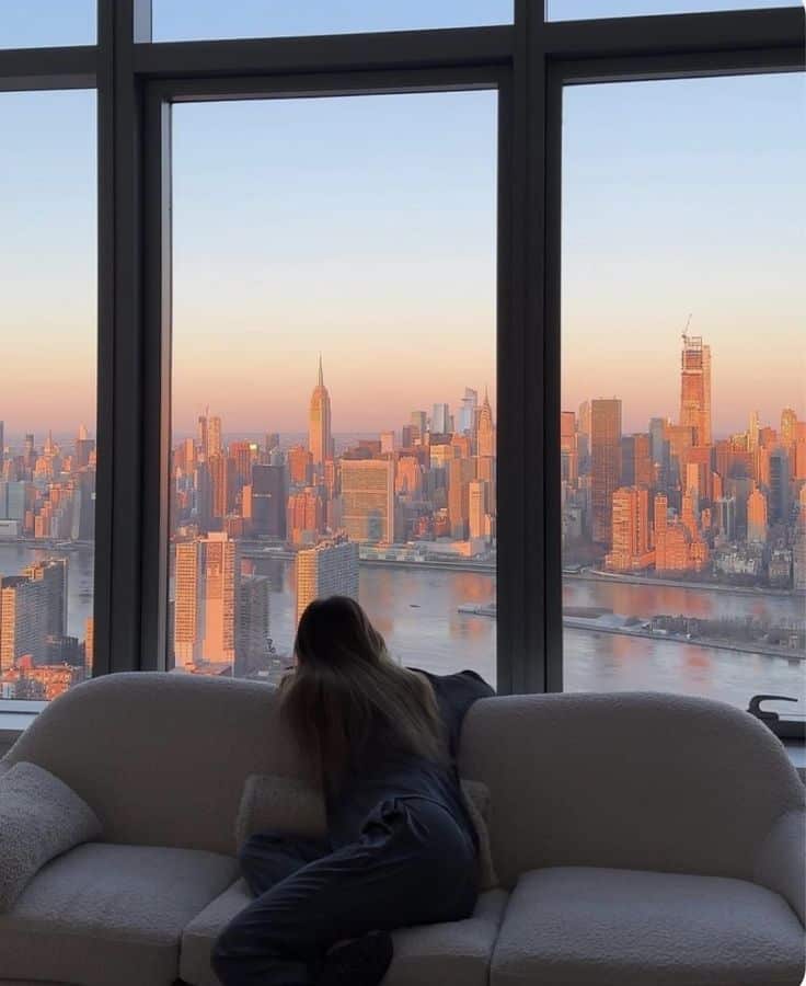 Woman gazing at New York City skyline with sunset; view from window capturing iconic buildings and vibrant sky.