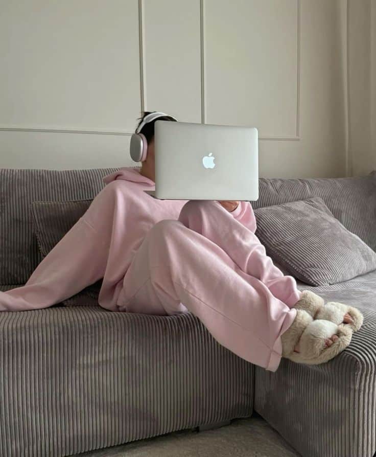 Person in pink loungewear on a couch using a laptop, wearing headphones and cozy slippers. Relaxed home office setting.