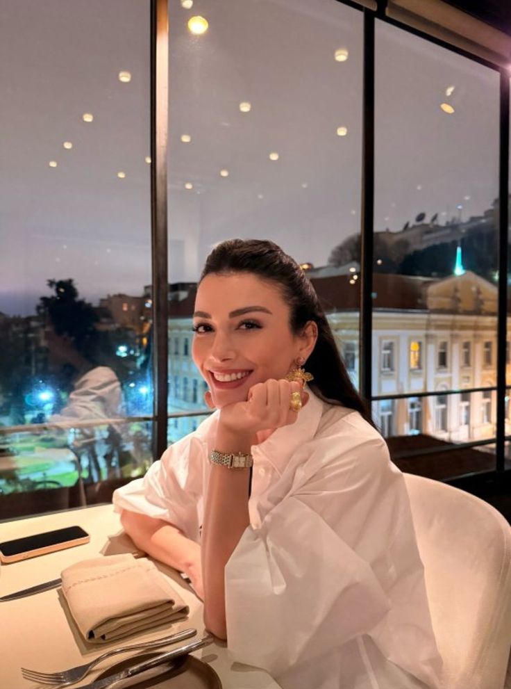 5704730e-fa5f-4f84-8fb1-4f74c18b0010 - Handy Home Smiling woman in a white blouse dining with a city view in the background, sitting at a restaurant table.