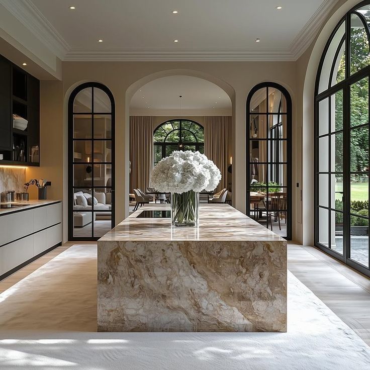 Luxurious kitchen with marble island, large windows, and elegant decor featuring a vase of white flowers. Modern and spacious design.