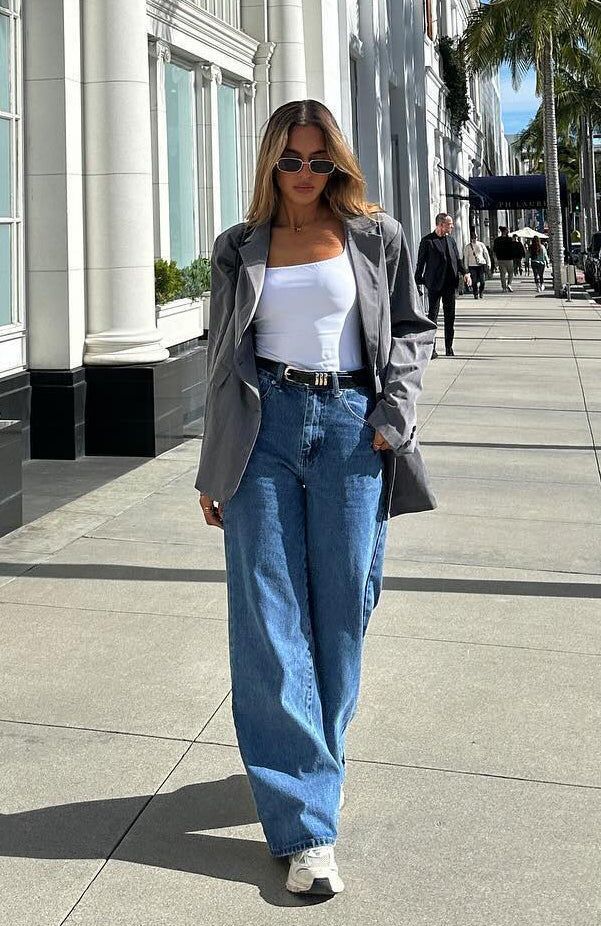Fashionable woman in a casual outfit: gray blazer, white tank, blue jeans, walking on a city sidewalk, sunglasses on a sunny day.