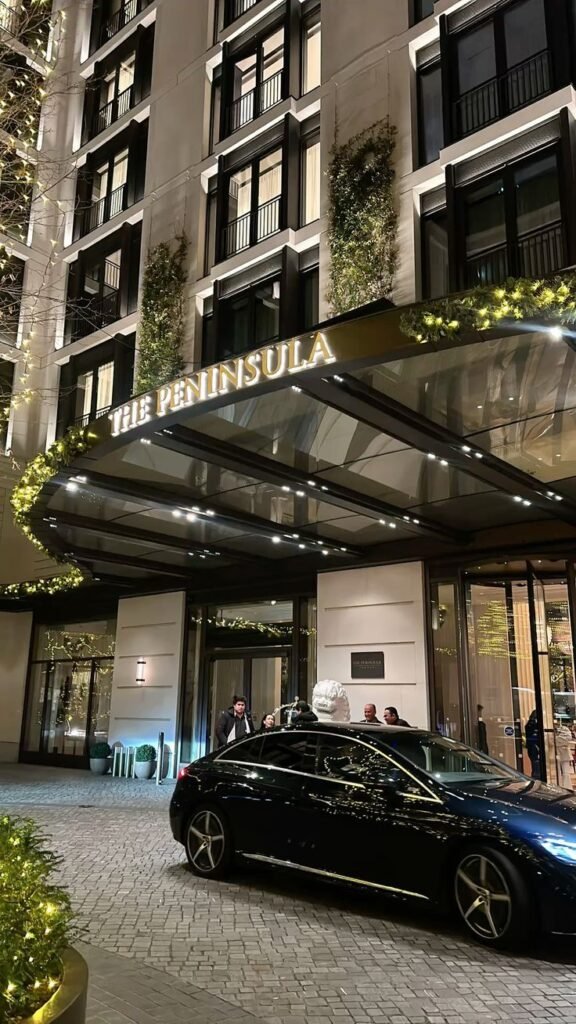 Entrance of The Peninsula Hotel adorned with festive lights, featuring a luxury car and glass canopy facade.