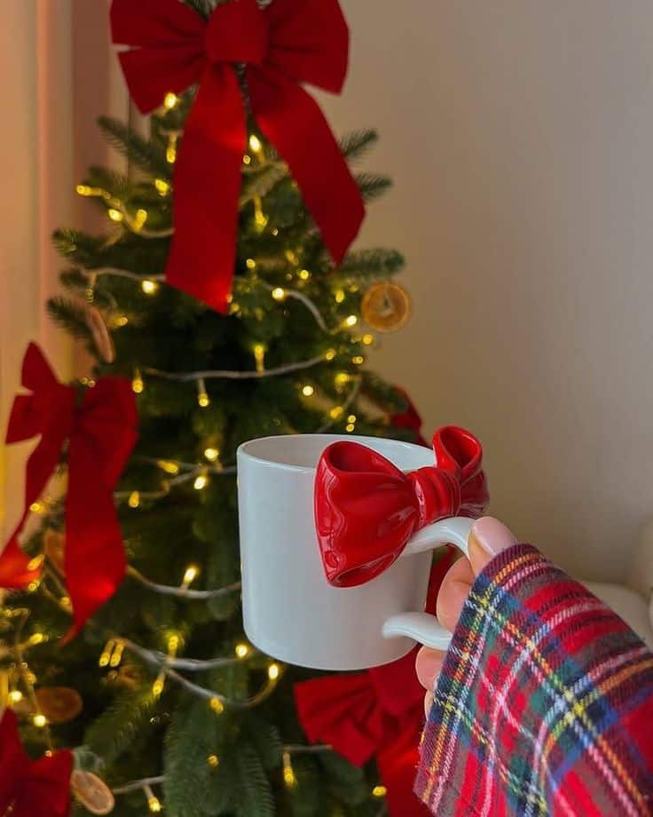 White mug with red bow, held by a plaid sleeve, in front of a decorated Christmas tree with lights and red bows. Festive ambiance.