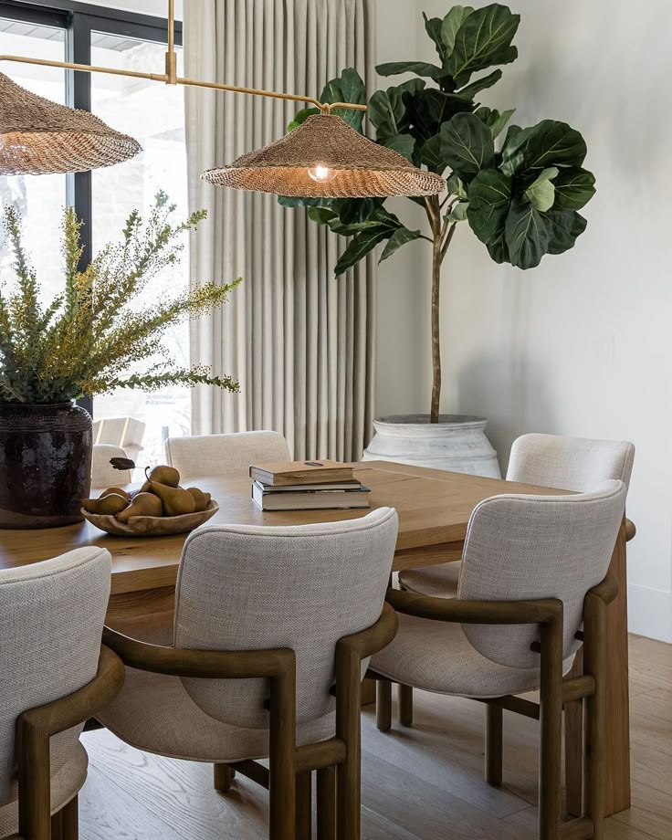 Modern dining room with a wooden table, beige chairs, woven pendant lights, and lush greenery for a stylish, earthy feel.