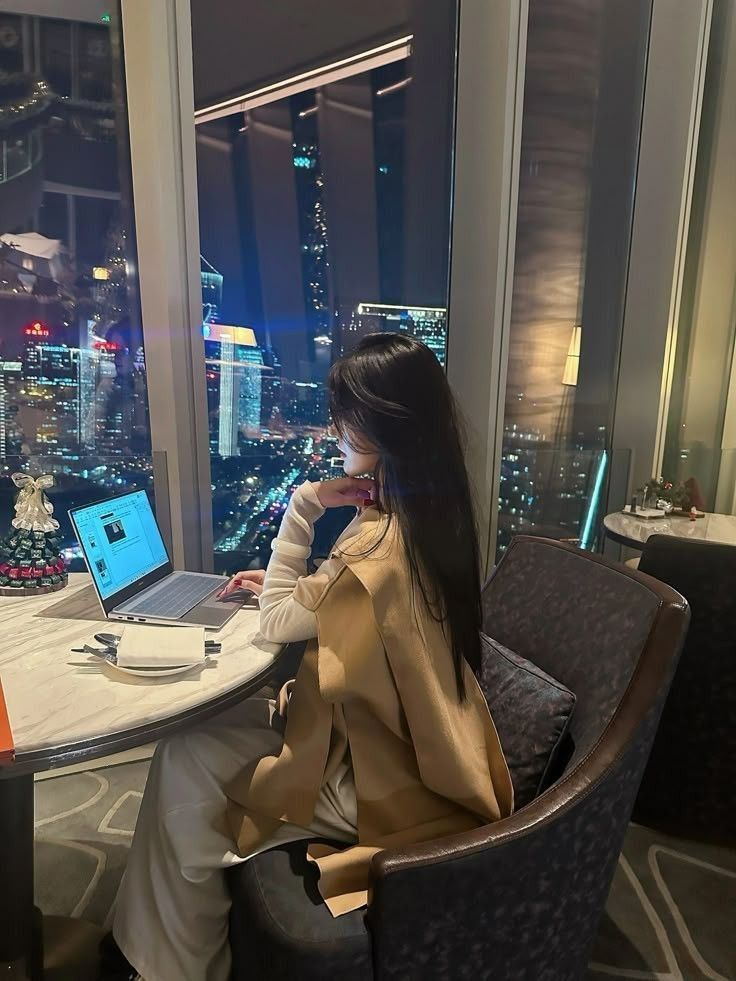 75cf6323-93ff-4993-98e2-4fe85072a9fb - Handy Home Person works on a laptop by a window with a night city view, dining table set nearby, suggesting a blend of work and relaxation.