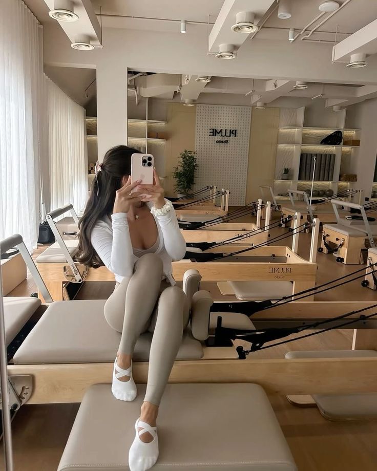 7e7e15a2-f21e-4e5a-b854-0abbf219eb01 - Handy Home Person sitting on a Pilates reformer machine in a modern studio, taking a selfie with a smartphone.