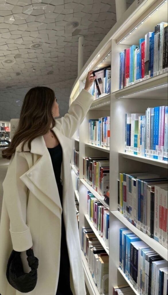 7ff30fd3-1849-4f4b-904f-81c1eba0976d - Handy Home Woman in a white coat browsing books at a modern library with white shelves and a patterned ceiling.