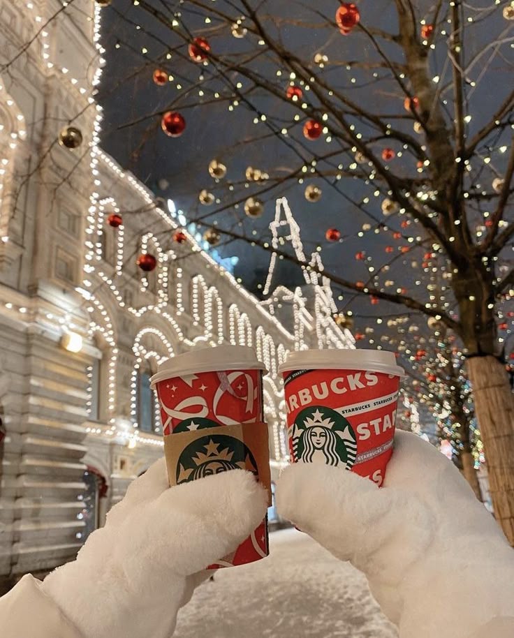 Festive holiday scene with hands in white gloves holding Starbucks cups, surrounded by twinkling lights and decorated tree.