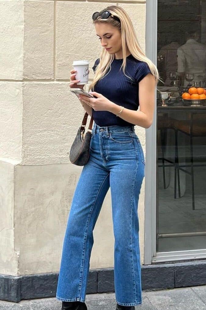 Stylish woman in jeans and boots holds coffee and phone, standing by a cafe. Trendy urban lifestyle.