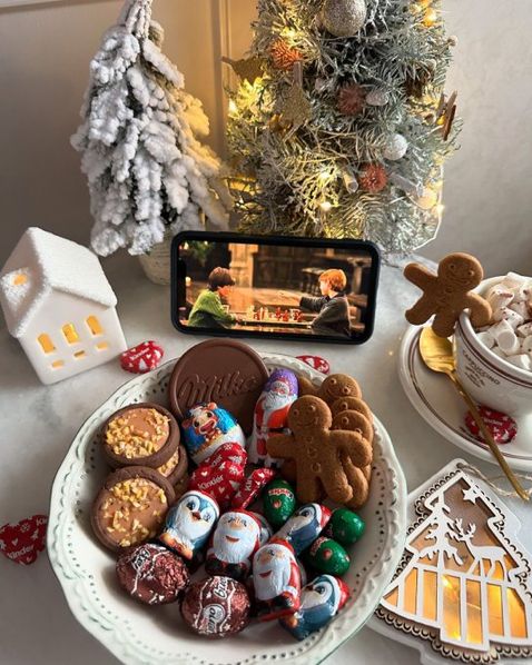 89198e9d-11fe-4a03-844f-4709fef23696 - Handy Home Cozy Christmas scene with chocolates, gingerbread, hot cocoa, phone streaming movie, and decorated tree in snowy setting.