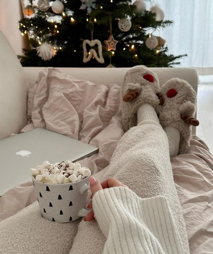 895e797b-ccee-4552-b940-521072c59d8f - Handy Home Cozy winter scene with person in fluffy slippers, hot cocoa, and a decorated Christmas tree, all beside a laptop on a sofa.