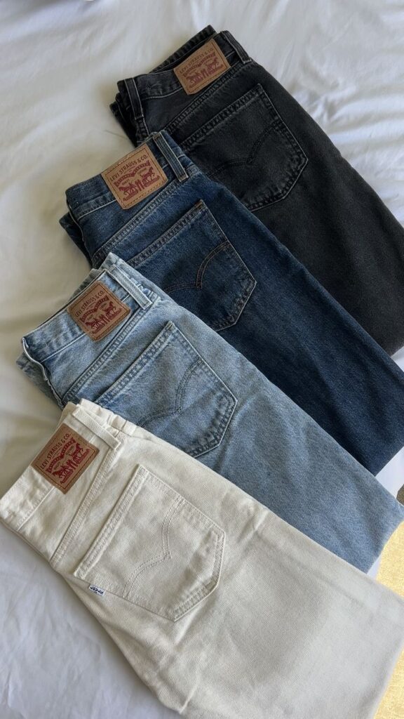 Stack of four Levi's jeans in black, dark blue, light blue, and cream on a white surface. Vintage style denim pants.
