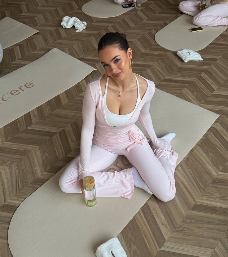900f1cb2-5ee9-401d-86c3-4dc214dffae4 - Handy Home Woman in pink yoga attire sitting on a mat, smiling with a water bottle indoors. Relaxed fitness setting on wooden floor.