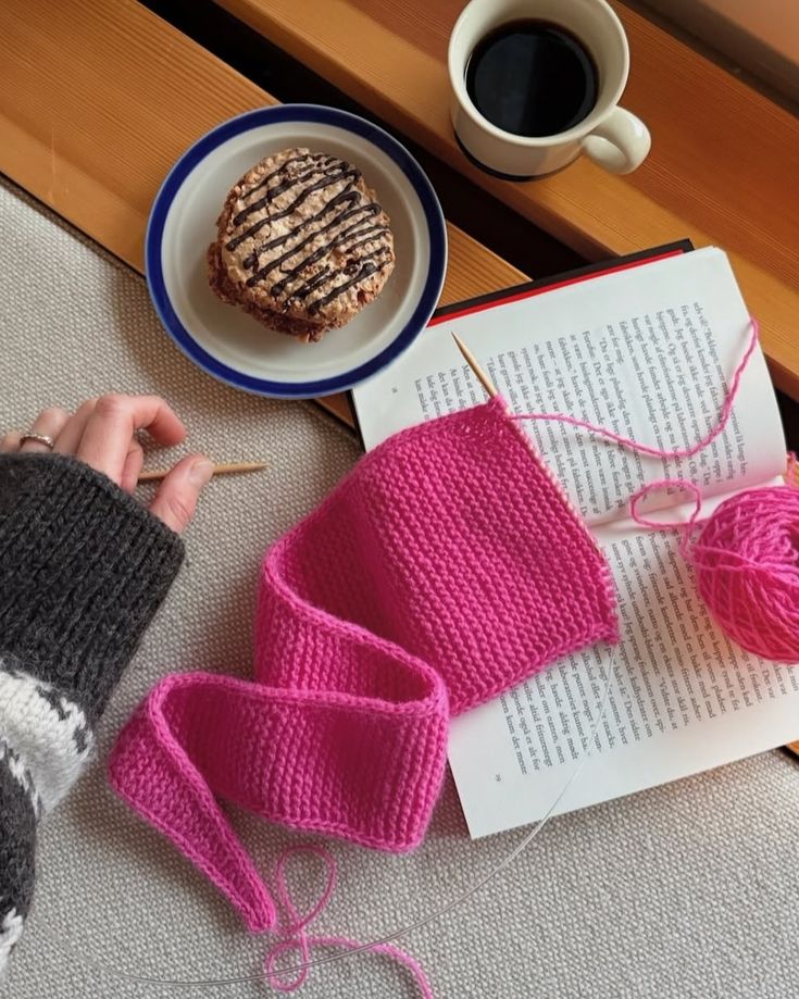Cozy knitting setup with pink yarn, coffee, an open book, and a pastry. Perfect for a relaxing day indoors.