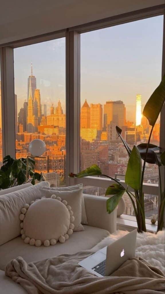 Cozy living room with city view at sunset; laptop on sofa, warm tones and plants enhance urban luxury.