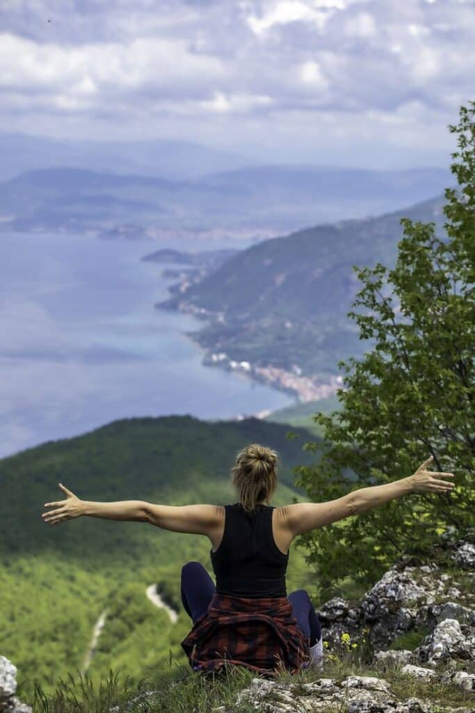 Best Things to do in Ohrid North Macedonia - Handy Home Woman enjoying scenic mountain view with open arms, overlooking a serene lake and green hills under a cloudy sky.