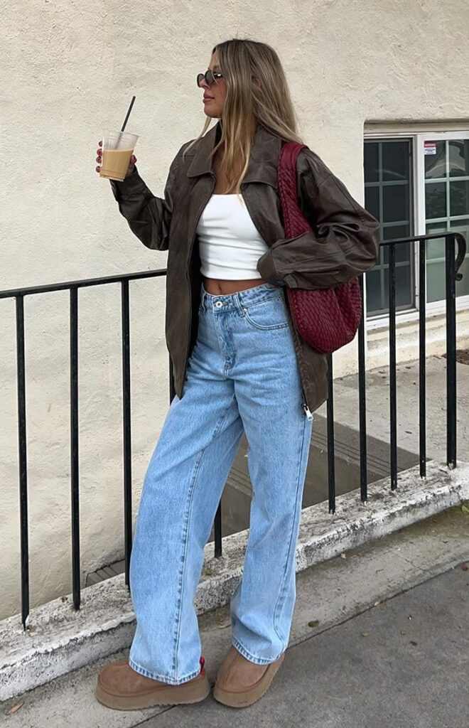 Woman in casual outfit with coffee, wearing a brown jacket, white top, blue jeans, and sunglasses, standing by a railing.
