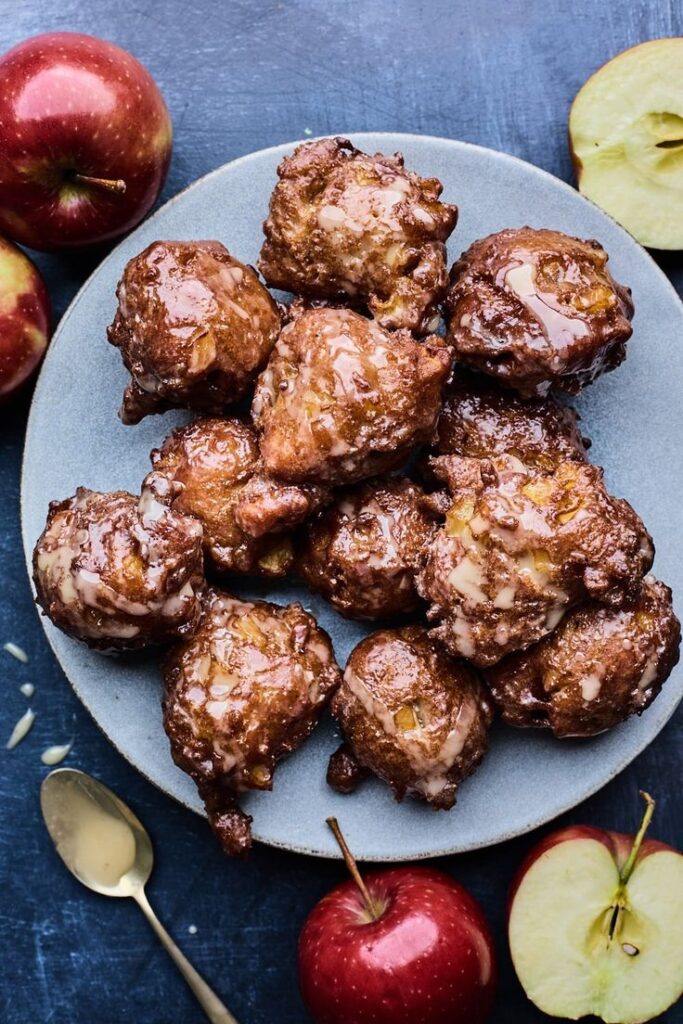 Chai Spiced Apple Cider Fritters With Maple Cider Glaze Olive Mango - Handy Home Plate of glazed apple fritters with fresh apples on a blue background, perfect for a sweet fall treat.