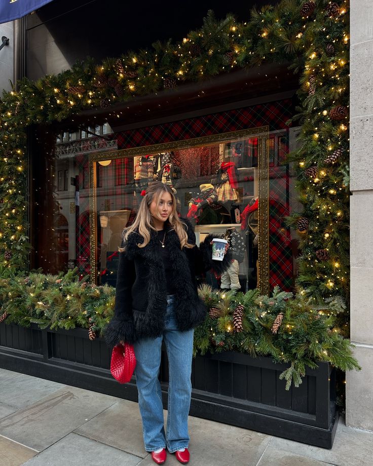 Christmas in London 🎄🎁 🎅🏼 - Handy Home Woman in winter fashion poses by a festive, decorated window display with lights and greenery. Cozy holiday shopping vibe.