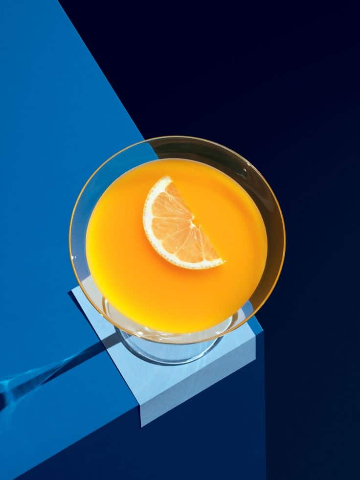 Complementary Colors Photography Tips - Handy Home Vibrant orange juice with a lemon slice in a glass, set against a bold blue geometric background.
