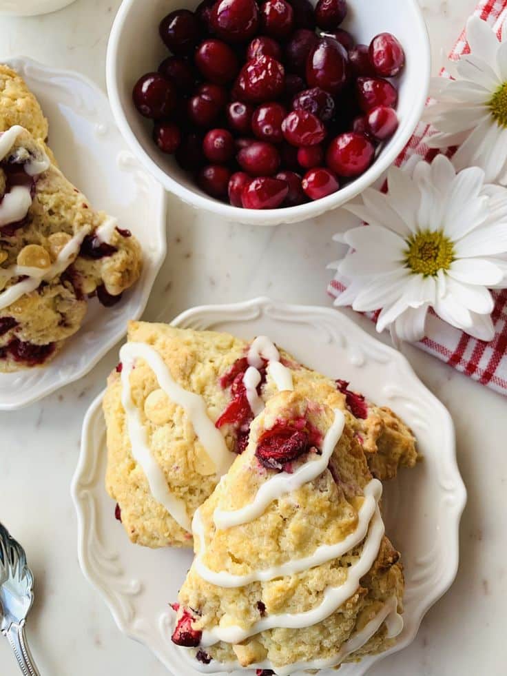 Cranberry White Chocolate Scones - Handy Home Cranberry scones with icing on a white plate, surrounded by fresh cranberries and white daisies, atop a checkered cloth.