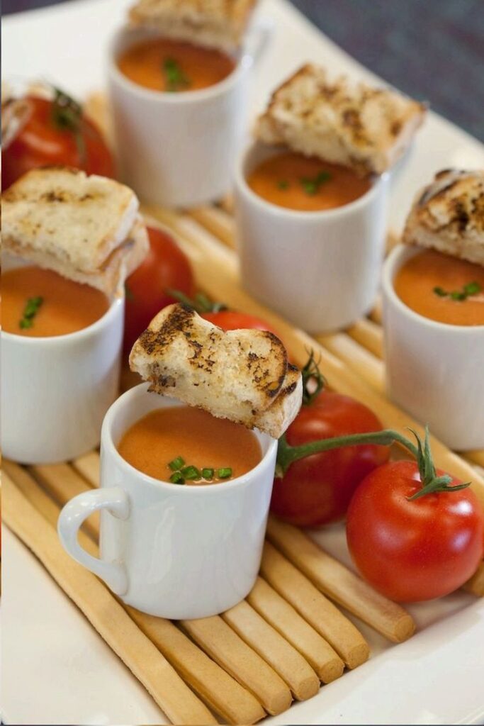 Delicious Mini Tomato Soup Shots with Grilled Bread and Breadsticks for Party Appetizers - Handy Home Mini mugs of tomato soup with grilled cheese and fresh tomatoes on a bamboo mat, perfect appetizer setup.