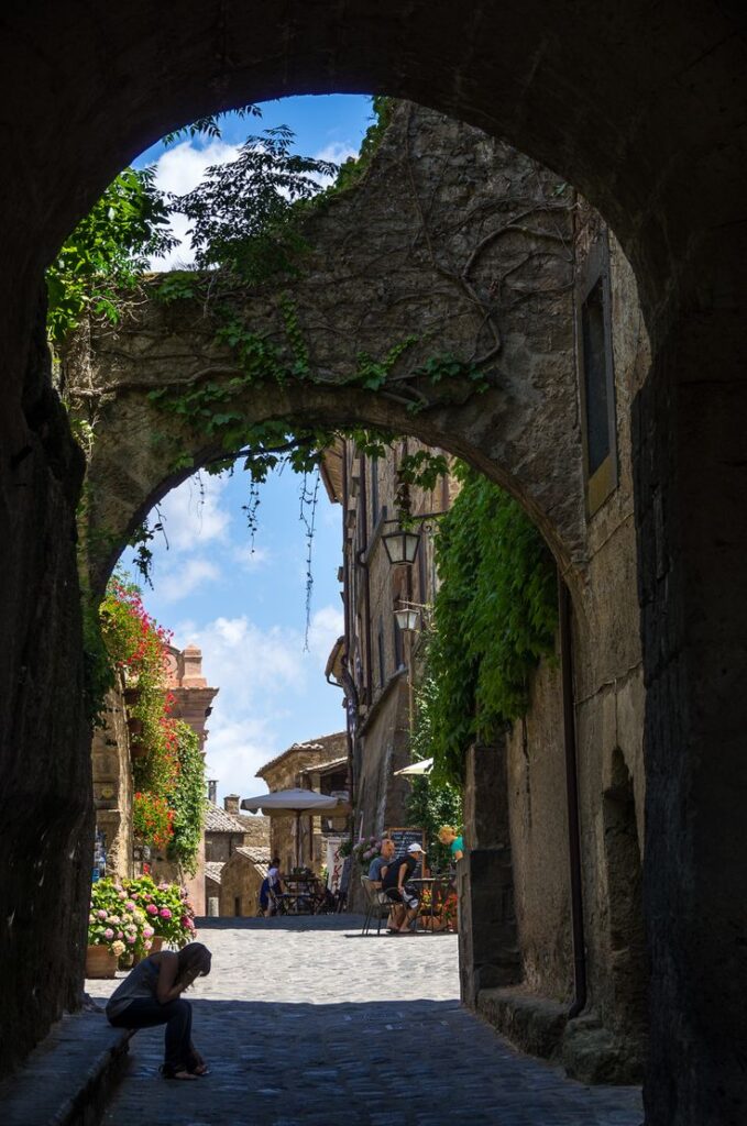 Entrance to Civita - Handy Home Charming cobblestone alley in a historic village, framed by an arch with lush greenery and vibrant flowers.
