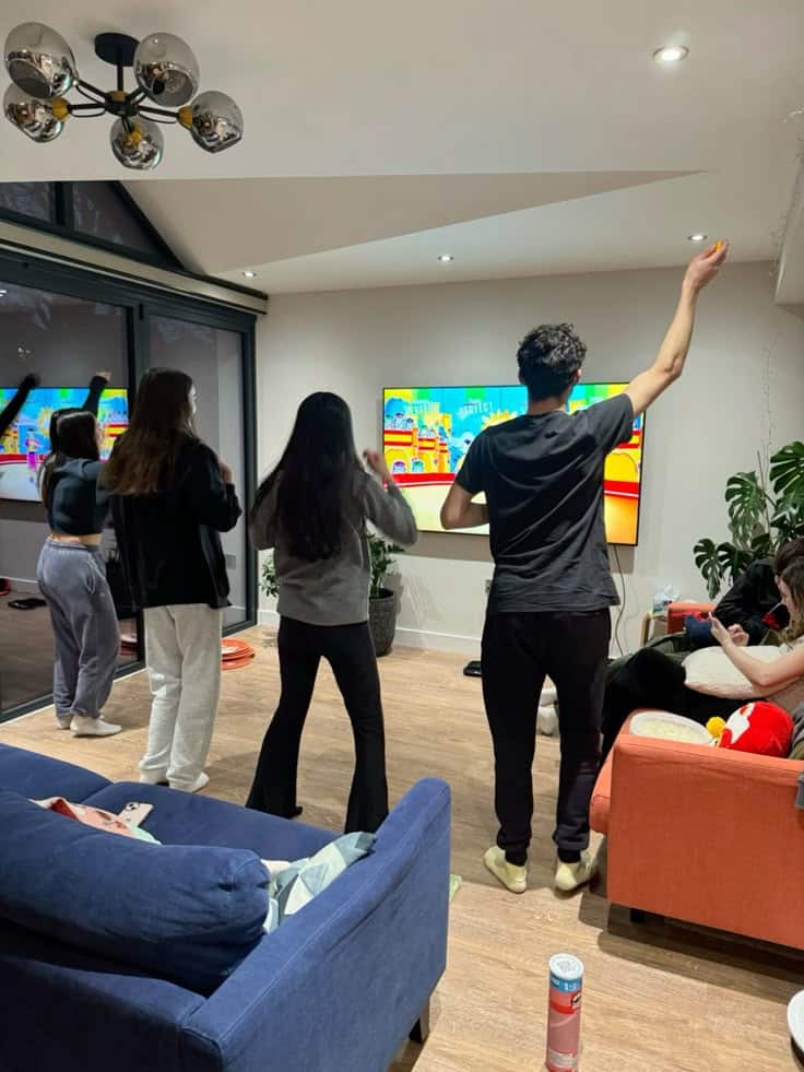 Group of friends playing a dance game on TV in a cozy living room with modern decor and snacks around.