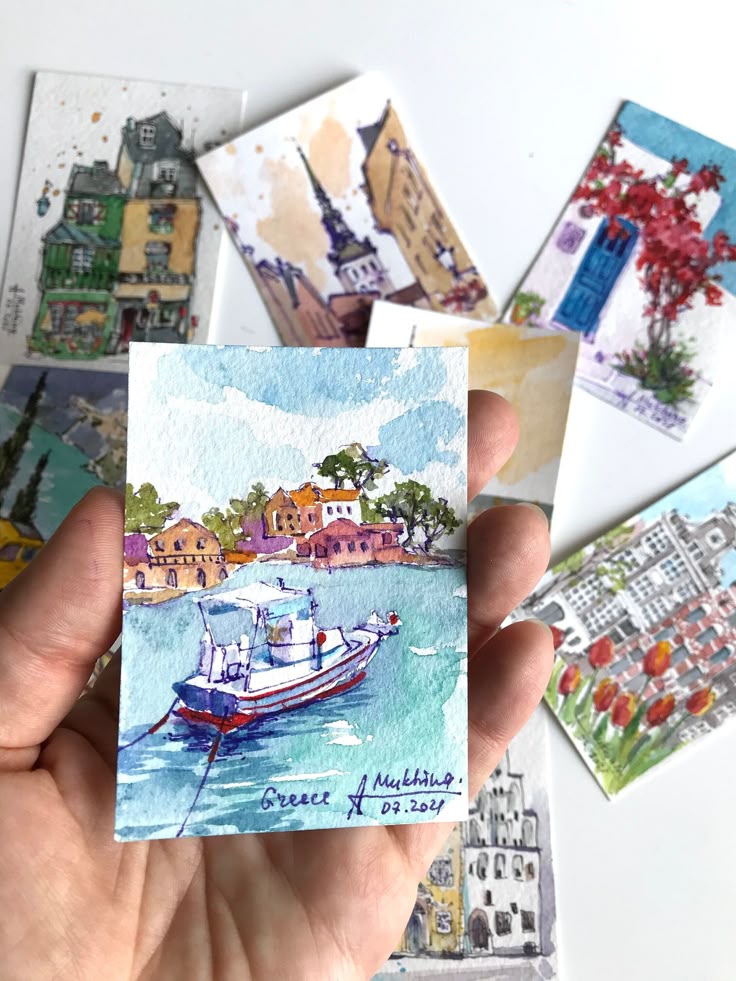 Hand holding colorful watercolor paintings of landscapes and architecture, featuring a boat scene titled Greece.