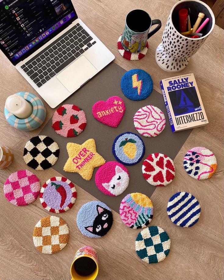 Colorful coasters on wooden table with laptop, book, and mugs; creative workspace decor and accessories.