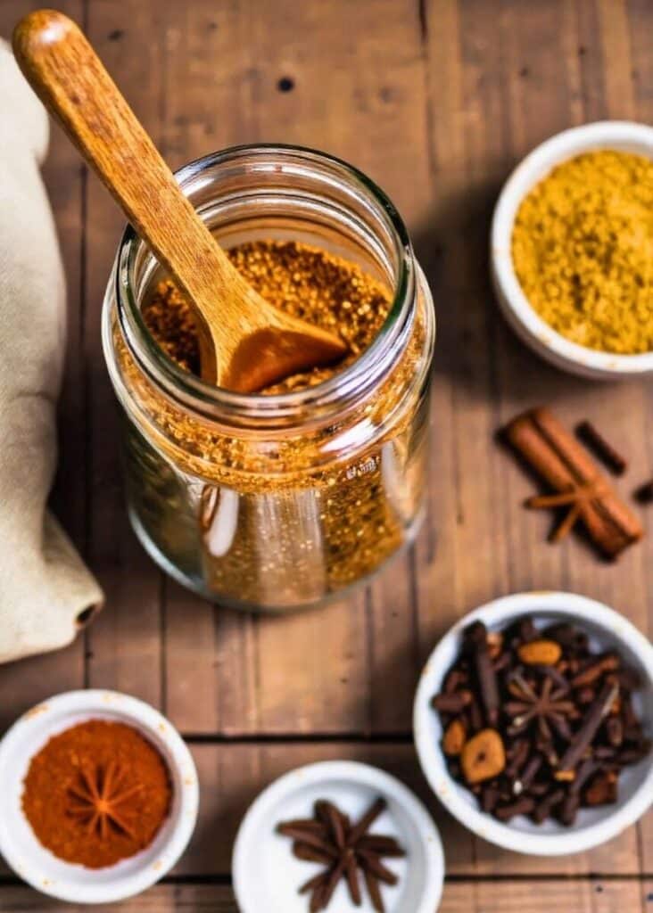 Jar of mixed spices with wooden spoon on wooden table, surrounded by bowls of various spices. Perfect for cooking inspiration.