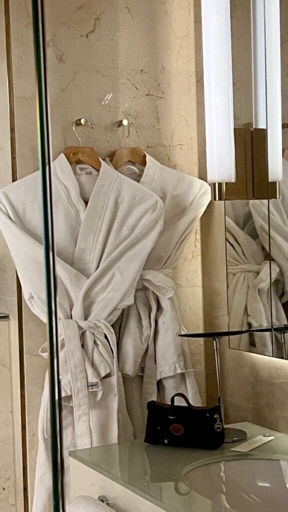 Two white bathrobes hanging in a luxurious bathroom with a small black handbag on the counter.