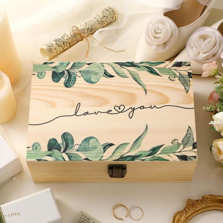 Wooden box with floral design and love you text, surrounded by wedding decor on table.