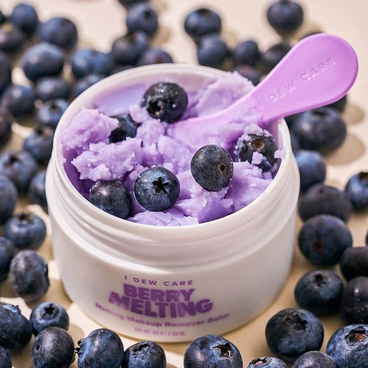 Purple makeup remover balm with blueberries and a pink spoon in a white container.