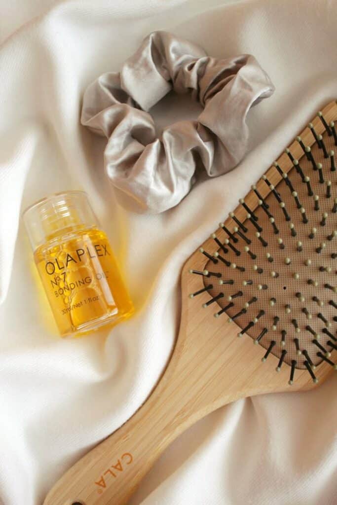 Instagram_ hiharleen - Handy Home Hair care essentials: bonding oil, wooden paddle brush, and silk scrunchie on a soft white fabric. Perfect for sleek styles.
