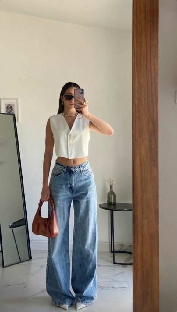 Woman in white sleeveless top and wide-leg jeans takes mirror selfie, holding a brown handbag in a stylish room. Fashionable look.