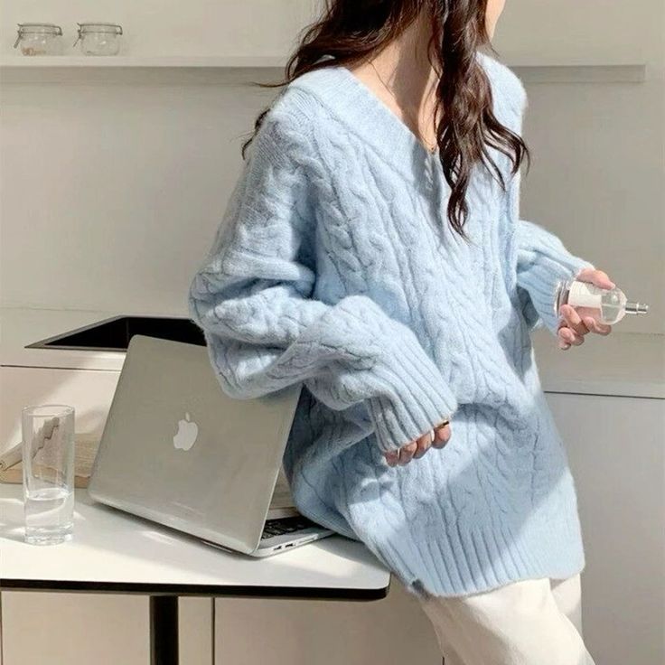 Woman in cozy blue sweater leaning on laptop, holding perfume bottle. Light, airy setting.